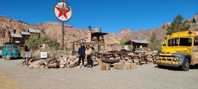 A couple poses at a Nevada Ghost Town near Las Vegas, surrounded by vintage cars, a Texaco sign, and desert mountains—ideal for ATV tours.