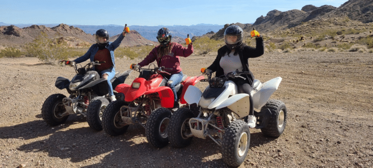 Explore Nevadas desert near the Colorado River on an ATV tour! Riders on black, red, and white ATVs give thumbs-up amidst rocky hills.