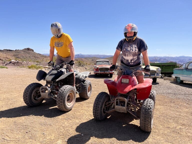 Explore Nevadas desert on an ATV tour! Ride quads—black and red—past vintage cars under a blue sky near the Colorado River, Las Vegas.