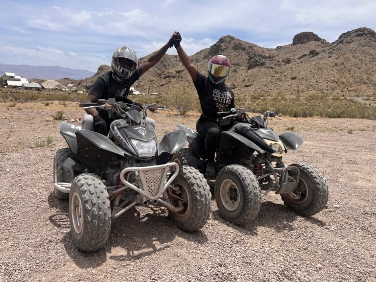 Two helmeted adventurers hold hands on ATVs in Nevadas desert landscape, near Las Vegas with mountains and a partly cloudy sky.