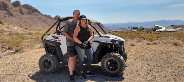 Couple smiles by a white off-road vehicle in Nevadas desert near the Colorado River. Explore ATV tours and ghost town sights!.