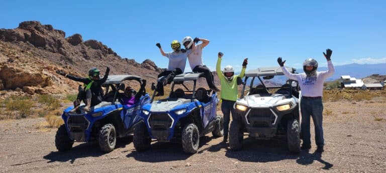 Adventure awaits near Las Vegas! Five pose with ATVs in Nevadas rocky desert, helmets on, under blue skies with distant mountains.