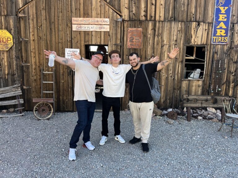 Three people joyfully pose with arms wide near a rustic, sign-adorned building in Nevada, perfect for Ghost Town sightseeing or ATV tours.