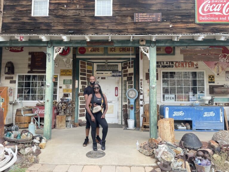 A smiling couple poses at a rustic Nevada store near the Colorado River, showcasing vintage Coca-Cola signs, perfect for Ghost Town sightseeing.