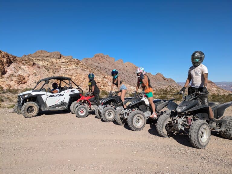 Four helmeted riders on ATVs line up beside an off-road vehicle in Nevadas rocky desert, perfect for Las Vegas RZR and ATV adventures.