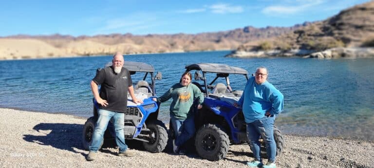 Three adventurers smile on a pebbled beach in Nevada, enjoying ATV tours by the Colorado River near Las Vegas and rocky hills.