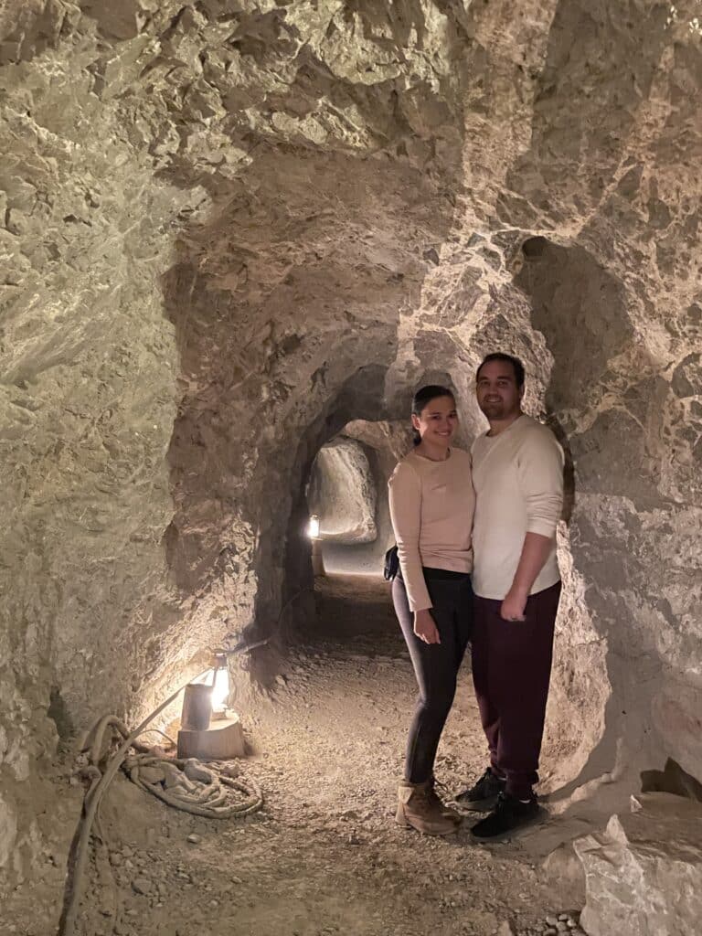 A couple stands in a dimly lit Nevada cave, part of their Colorado River adventure near Las Vegas, beaming with casual charm.