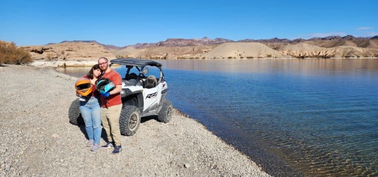 A couple on a Nevada lakeshore readies for a Las Vegas ATV Tour. The serene lake and off-road RZR hint at thrilling ghost town adventures.