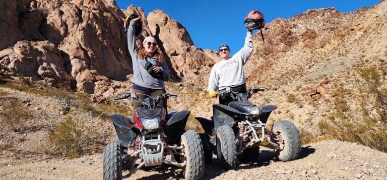 Two adventurers raise helmets by their ATVs on a rocky Nevada trail. Enjoy Las Vegas ATV tours with stunning views of rugged cliffs!.