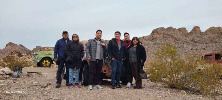 A group of six in winter jackets stands by a rusty car in rocky Nevada terrain, set for a thrilling Las Vegas ATV tour near the Colorado River.