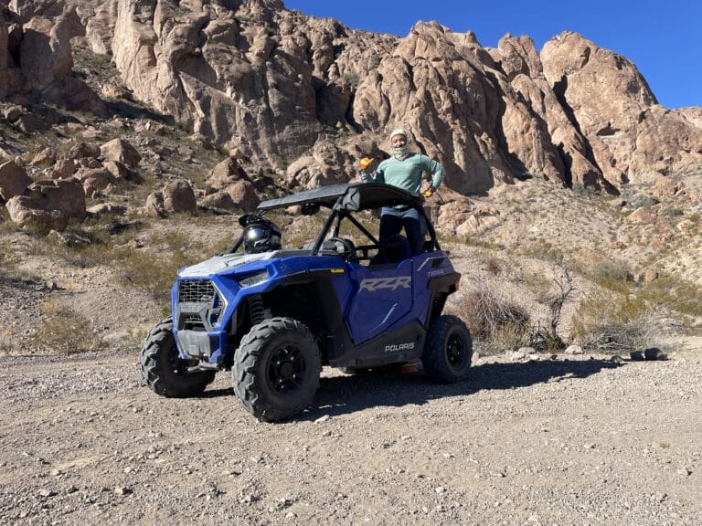 Explore thrilling ATV tours in Nevada! Near the Colorado River, a man in sunglasses stands by a blue Polaris RZR amid Las Vegass rocky terrain.