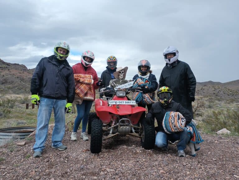 Explore Nevada with Las Vegas ATV tours! Six adventurers in helmets pose by a red ATV, ready for RZR off-road rides and Ghost Town sightseeing.