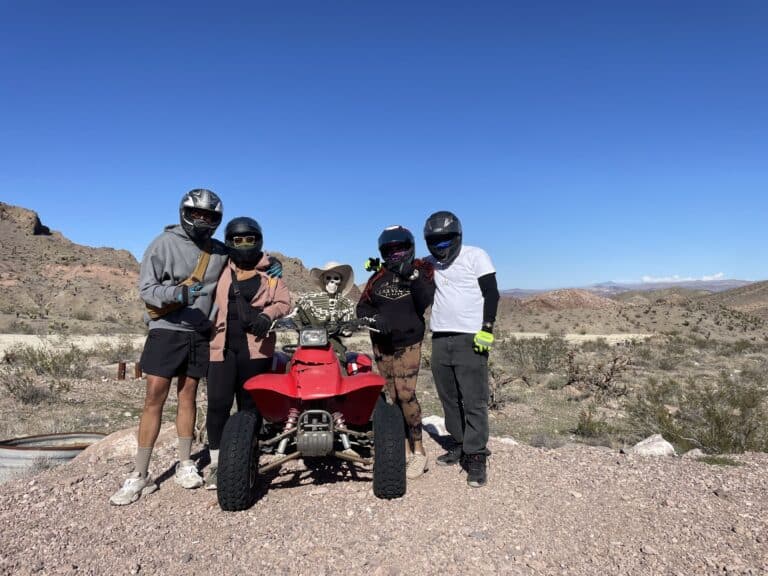 Four helmeted adventurers pose with a red ATV and skeleton in Nevadas desert. Explore thrilling Las Vegas ATV tours and RZR rides.