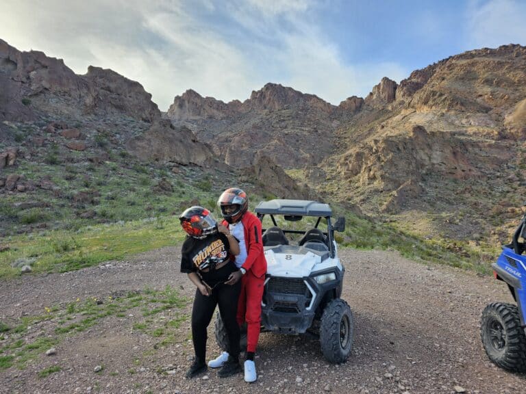 Two helmeted adventurers, one in black and the other in red, stand by an ATV near a blue vehicle with Nevadas desert and mountains behind them.