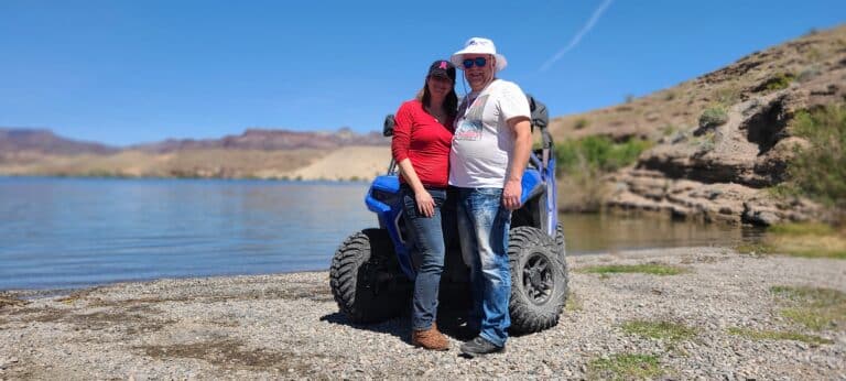 A couple embraces the thrill of RZR off-road rides by a blue vehicle on Nevadas rocky lakeshore, near the Colorado River.