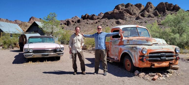 Two men smile by a vintage Dodge truck and pink car in Nevadas desert near Las Vegas, ideal for Ghost Town sightseeing and ATV tours.