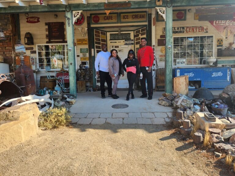 Four people smile by a rustic shop near the Colorado River, hinting at Historic Gold Mine Tours. Explore Nevadas ATV rides and ghost towns!.
