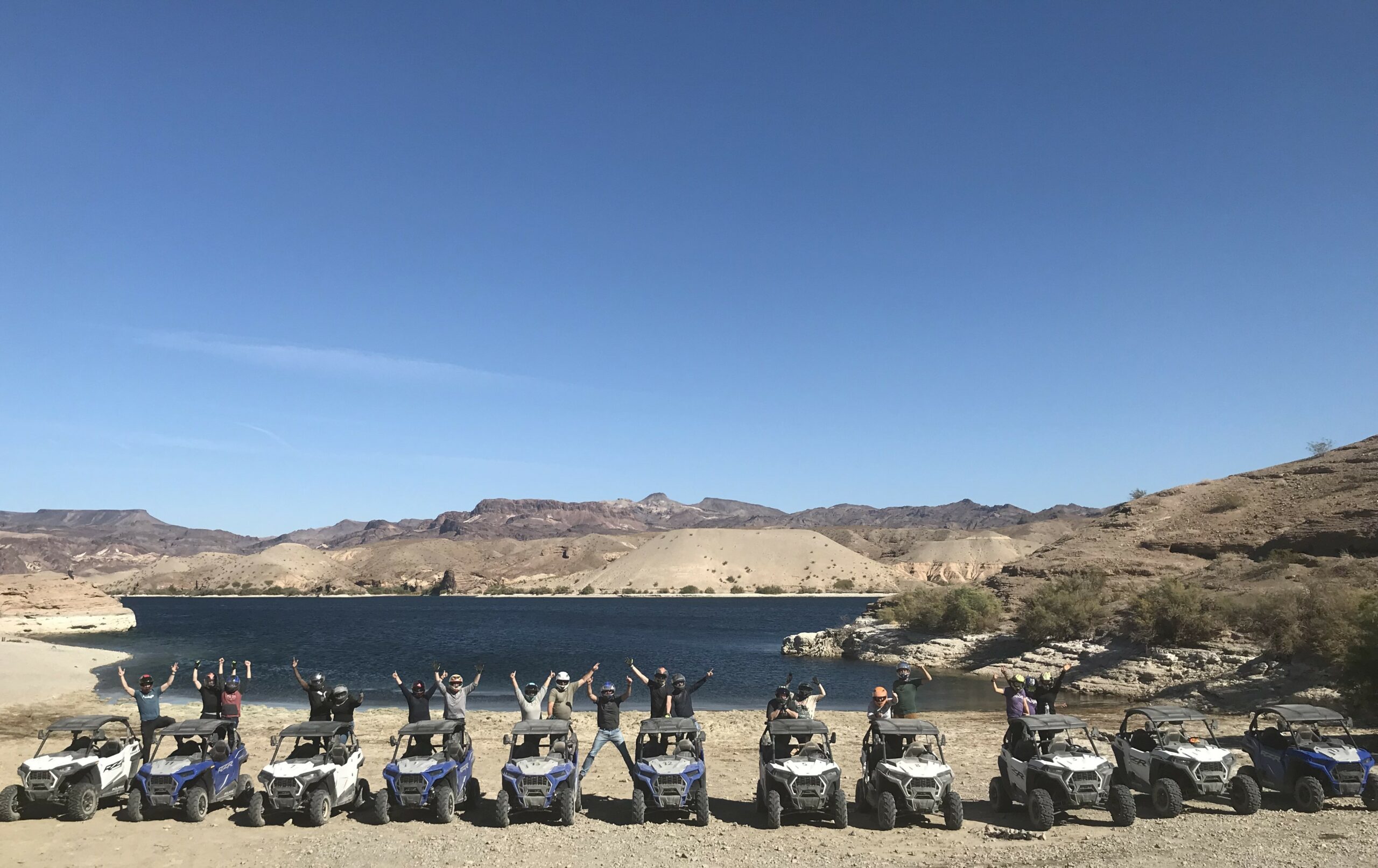 Group celebrates by off-road vehicles near a lake, perfect for Nevada ATV tours. Explore Las Vegas’ arid hills under clear skies!.