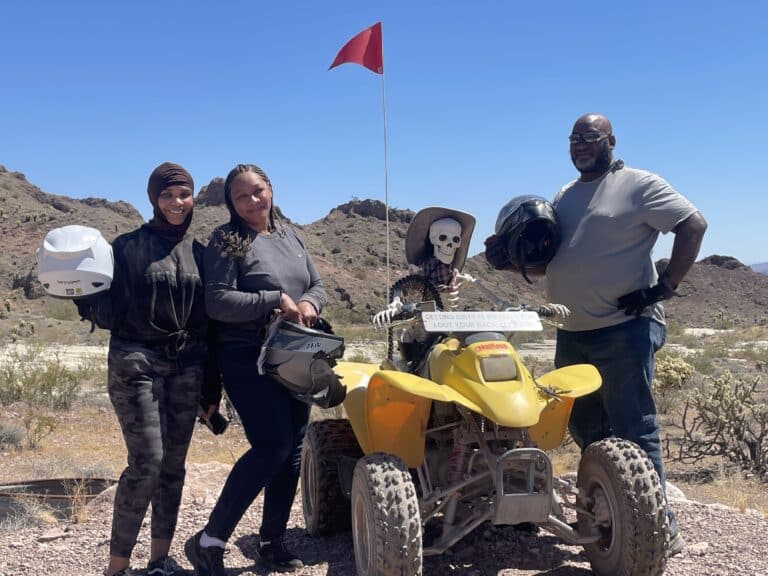 Four friends relish a Nevada adventure, posing with a yellow ATV by the Colorado River, ready for an exhilarating Las Vegas Ghost Town sightseeing tour.