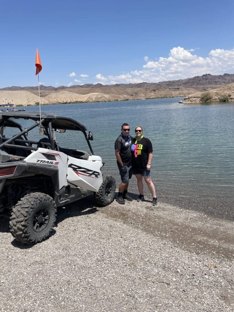 A couple gears up for an RZR off-road adventure by a tranquil Nevada lakeshore near Las Vegas, with mountains and the Colorado River as their backdrop.
