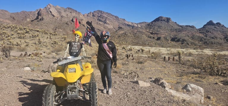 A helmeted person in a black jacket poses in Nevadas desert beside a yellow ATV near Las Vegas, with a skeleton rider. Perfect for thrilling ATV tours!.