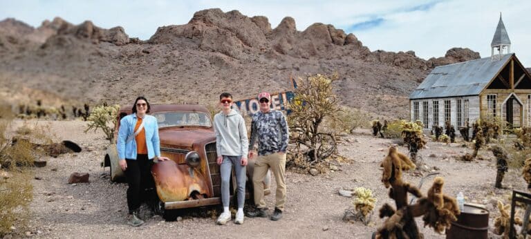 Three people in a Nevada desert near Las Vegas stand by a rusty car. Nearby, a chapel and Motel sign, with cacti set against rocky hills.