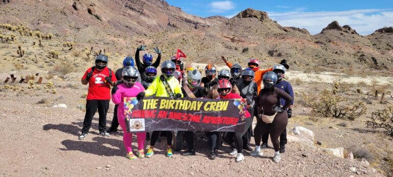 Adventure-seekers in helmets and vibrant gear pose with The Birthday Crew banner in Nevadas rocky desert near the Colorado River.