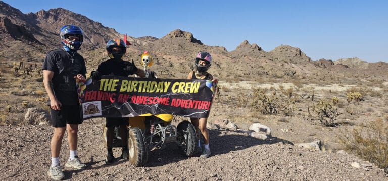Four adventurers in helmets celebrate with a Birthday Crew banner on an ATV tour near the Colorado River, Nevada. #LasVegasAdventure.