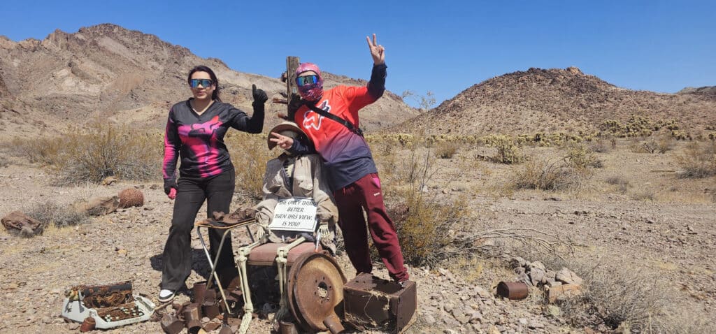 Three pose near an old wheelchair in Nevadas rocky desert. Enjoy the view sign. Hills & sparse plants backdrop, near Las Vegas ATV tours.