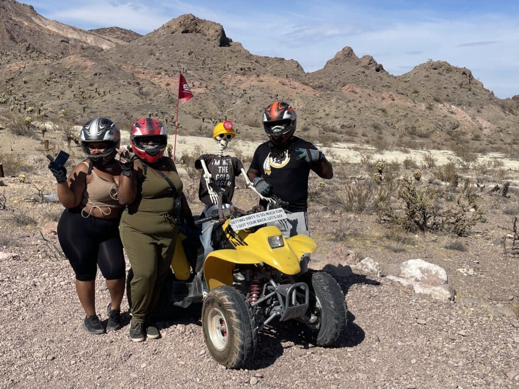 Four adventurers with helmets pose by an ATV in Nevadas rocky desert near the Colorado River, enjoying a sunny Las Vegas off-road tour.