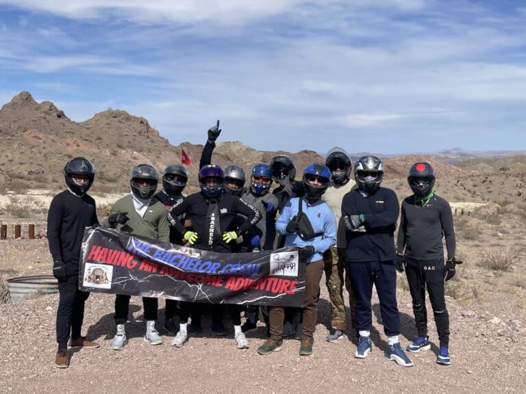 A group of 10, geared up for adventure in Nevada, poses on rocky terrain with a banner: THE BACHELOR CREW HAVING AN AWESOME ADVENTURE. Perfect for Las Vegas ATV tours or Colorado River RZR off-road rides.