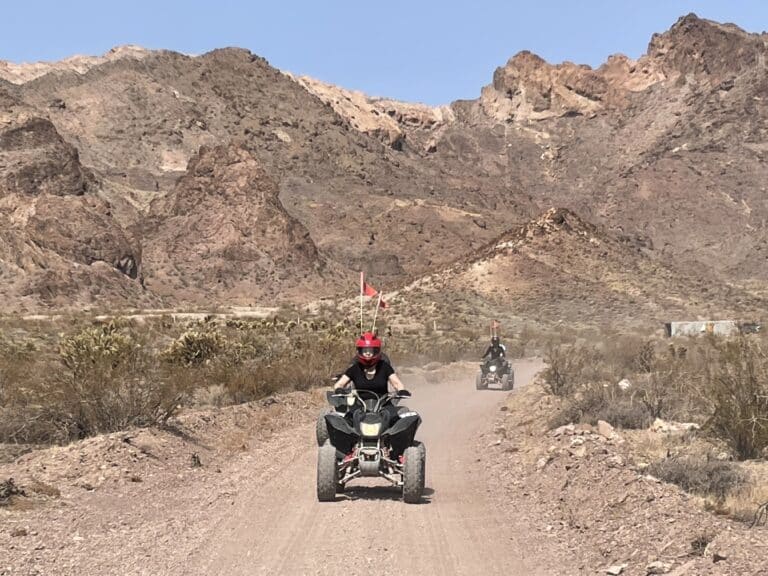 Explore the Nevada desert near Las Vegas on an ATV tour. Riders traverse rocky trails past craggy hills and sparse greenery under blue skies.