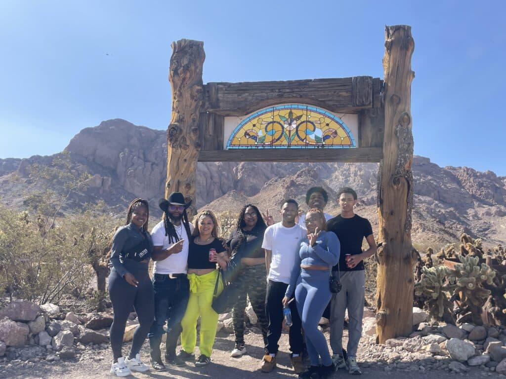 Eight friends smile by a wooden structure with stained glass, set against Nevadas desert and mountains near Las Vegas. Perfect for ATV adventures!.