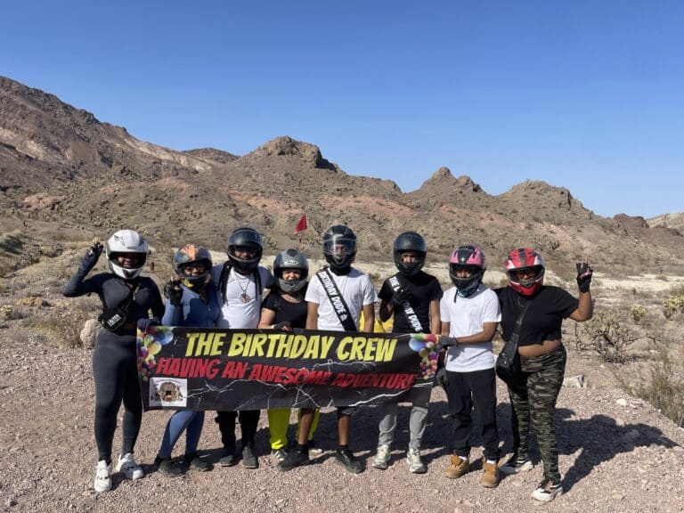Nine friends in helmets pose with a BIRTHDAY CREW ADVENTURE banner during an ATV tour near Las Vegas, against Nevadas rocky landscapes.