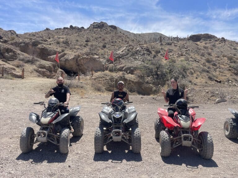 Three friends enjoy an ATV tour near Las Vegas, Nevada, riding RZRs with red flags in a sunny desert by rocky hills.