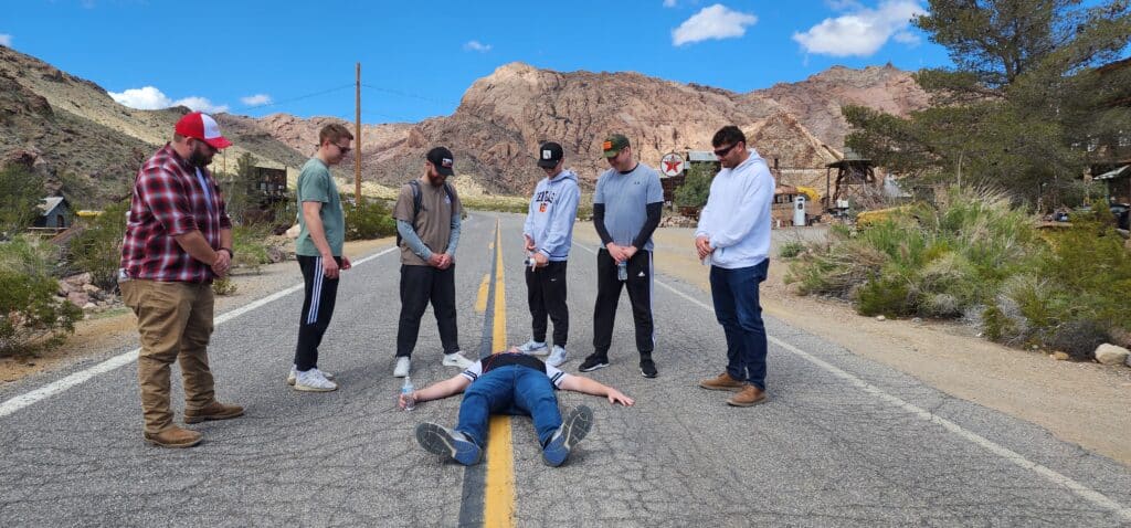 Six men on a Ghost Town ATV Tour in Las Vegas attend to another man on a rocky Nevada road, beneath a blue sky.