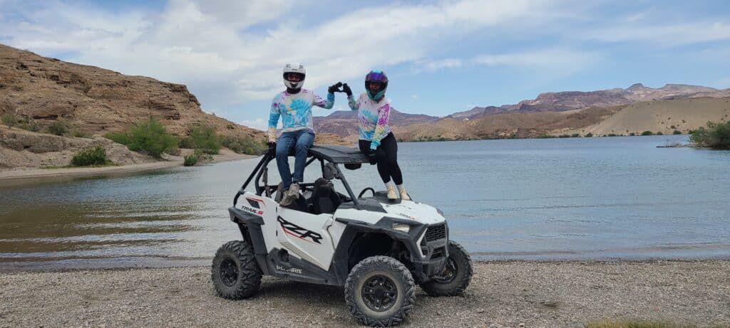 Two adventurers in helmets and tie-dye shirts pose on an RZR in Nevada near the Colorado River. Experience thrilling Las Vegas ATV tours!.