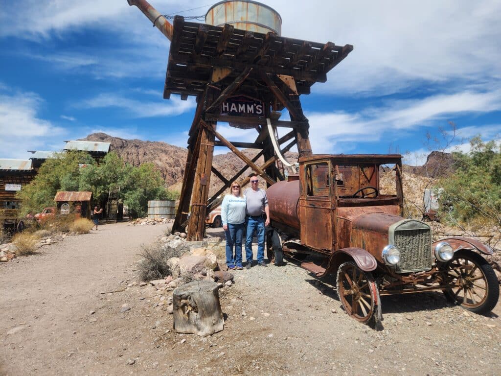 Smiling duo at Hamm’s water tower & vintage truck in a Nevada desert ghost town near Las Vegas—perfect for ATV or RZR adventures!.
