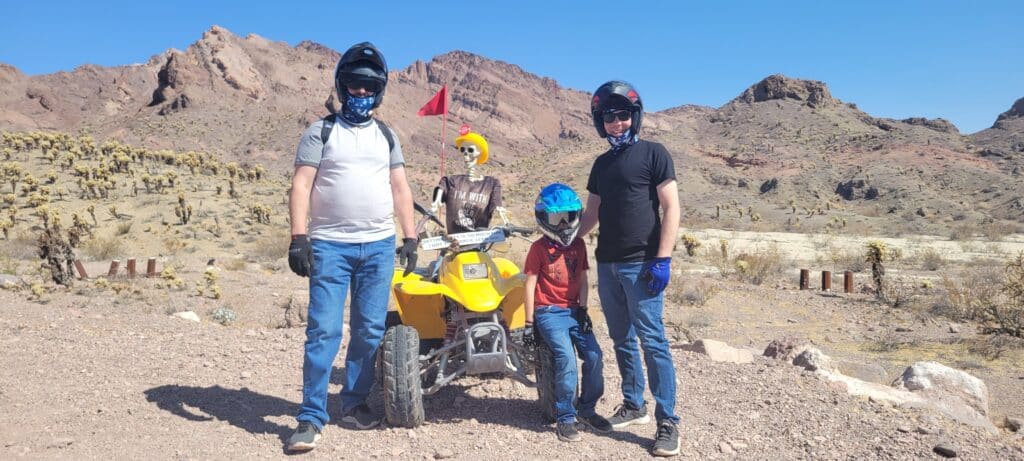 Riders in helmets pose by a yellow ATV near Las Vegas, Nevada; a skeleton mannequin joins this fun desert ATV tour adventure.