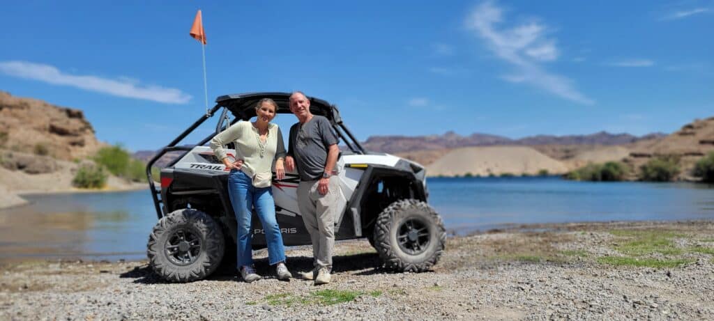 Smiling couple poses by a white RZR on rocky Nevada terrain near the Colorado River, enjoying an epic Las Vegas ATV tour adventure.