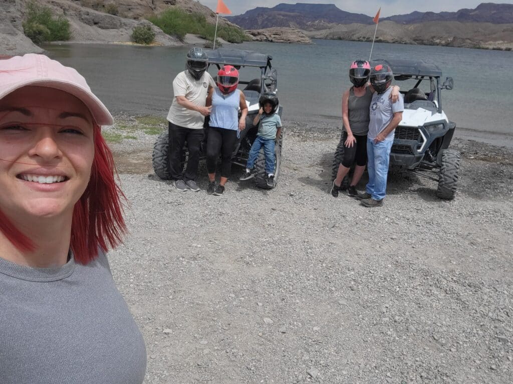 Red-haired woman takes a selfie at Nevadas Colorado River as group enjoys RZR ATV tour near Las Vegas with scenic hills and water.