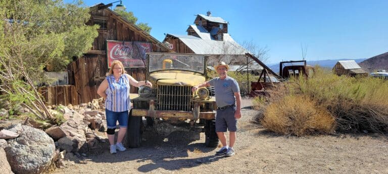 Two people pose by a vintage yellow jeep near rustic buildings and desert plants on a Nevada ghost town tour near Las Vegas.