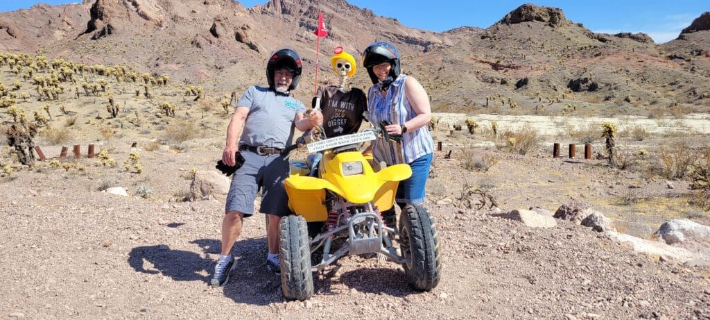 Smiling duo in helmets by a yellow ATV with an alien figure, exploring Nevada desert near Las Vegas on an exciting ATV tour.