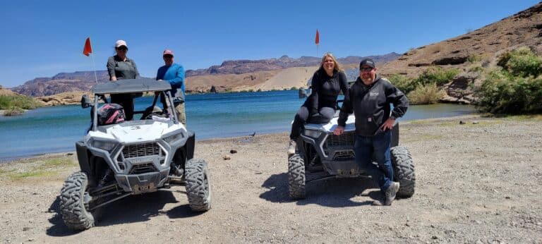 Friends enjoy an ATV tour near Las Vegas, posing by RZRs at a blue Colorado River lake with Nevada mountains behind.