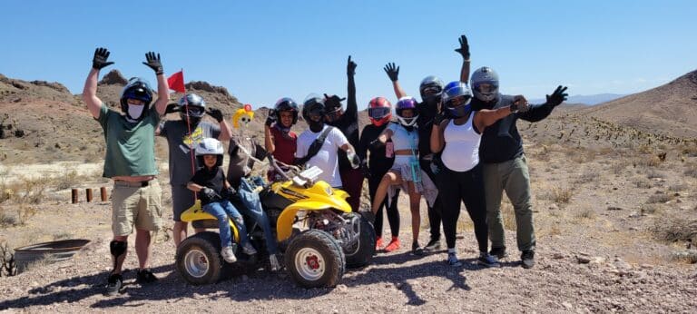 Adventure seekers in helmets pose by a yellow ATV in the Nevada desert near Las Vegas with mountains and blue skies—perfect for ATV tours!.