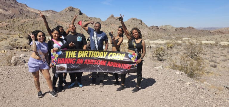 Eight women pose in the Nevada desert near Las Vegas, holding a Birthday Crew banner after a Colorado River ATV adventure.
