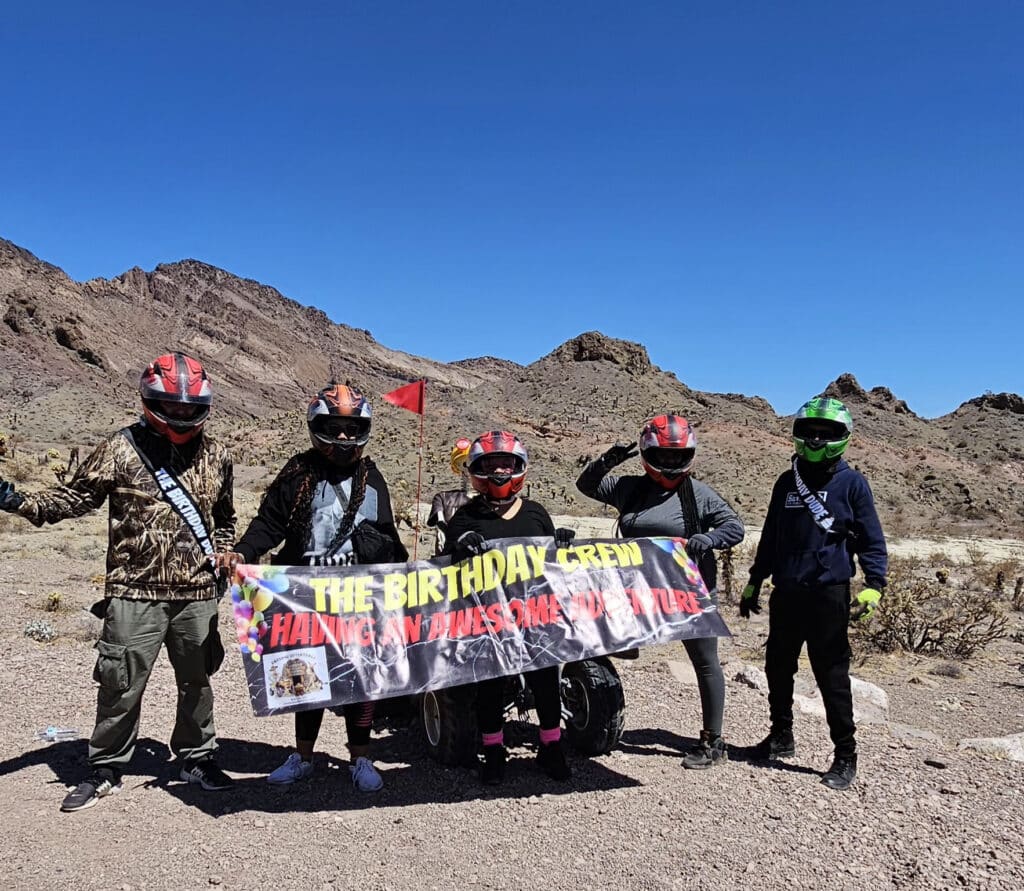 Five adventurers in helmets celebrate with a banner during a Las Vegas ATV tour by the Colorado River in Nevadas rocky desert.
