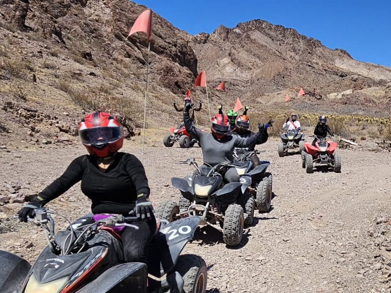 Thrill-seekers ride ATVs on a Nevada desert trail near Las Vegas, with red flags waving and mountains by the Colorado River.