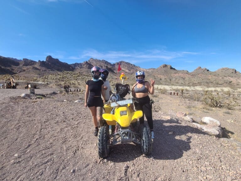 Four riders in helmets pose by a yellow ATV near Las Vegas, Nevada, on an ATV tour with mountain views under a clear blue sky.