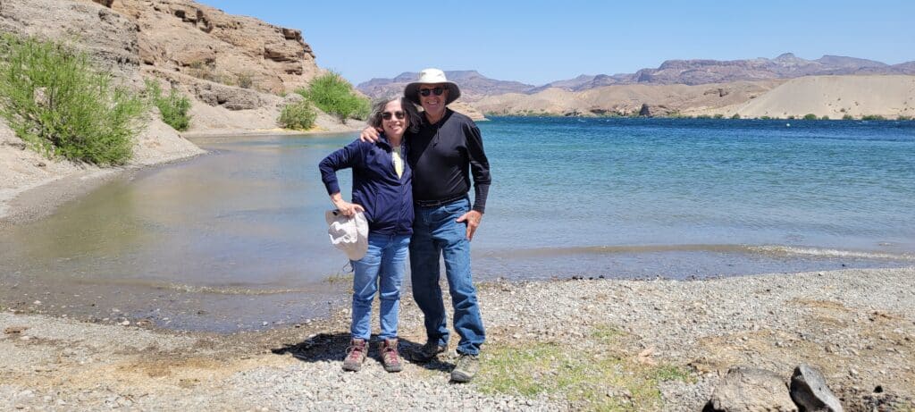 A smiling couple poses on a rocky Nevada lakeshore near the Colorado River, enjoying ATV tours with mountains and clear skies in view.